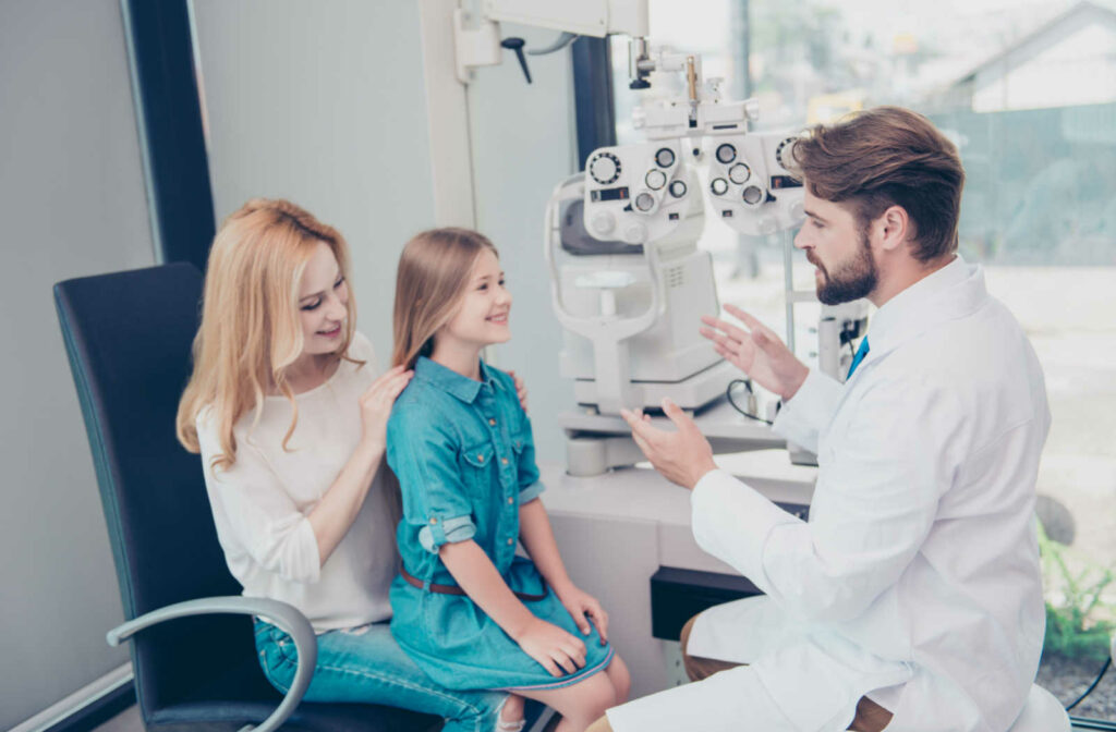 An optometrist talking to a young girl, who is sitting on her mother's lap about her upcoming eye exam.