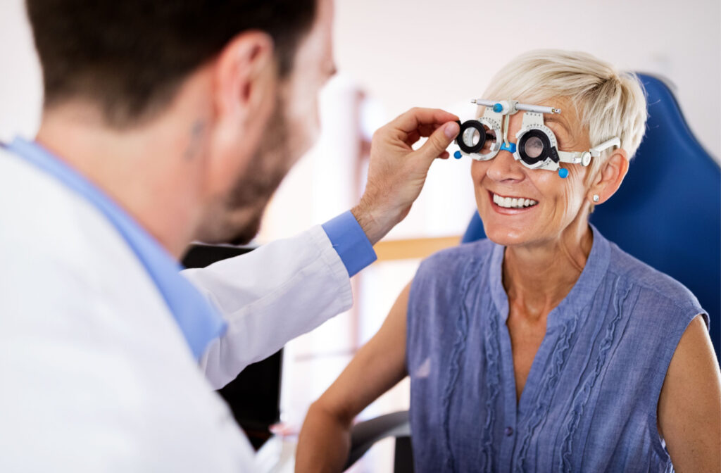 A smiling senior woman in a blue blouse is sitting in a chair while a young male optometrist examines her eyes.